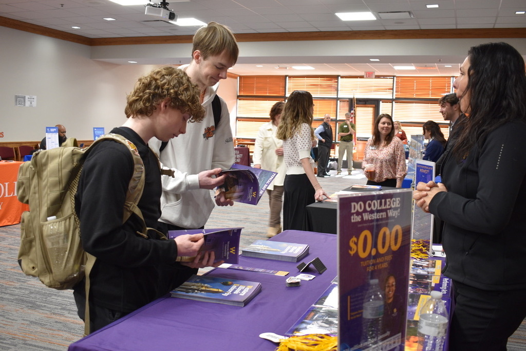 A group of people inside a room with tables and brochures. A boy is looking at a booklet.
