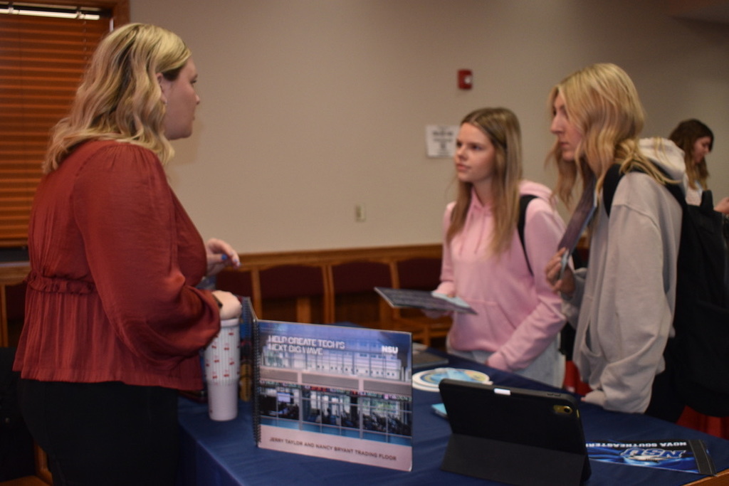 Three women in a room with a table displaying pamphlets. One woman holds a folder.