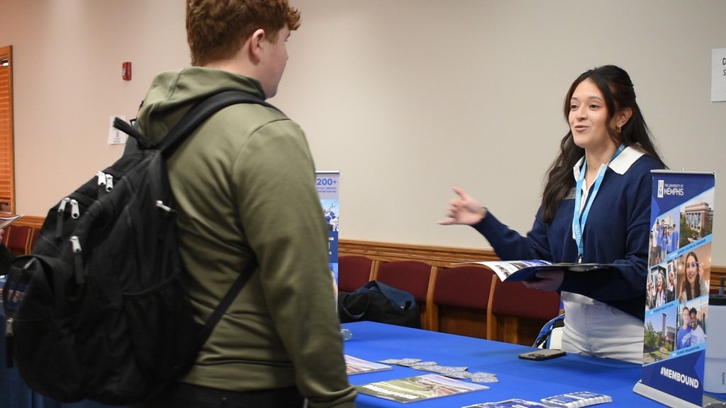 Two people are indoors, one with a backpack stands and speaks with another person at a table.