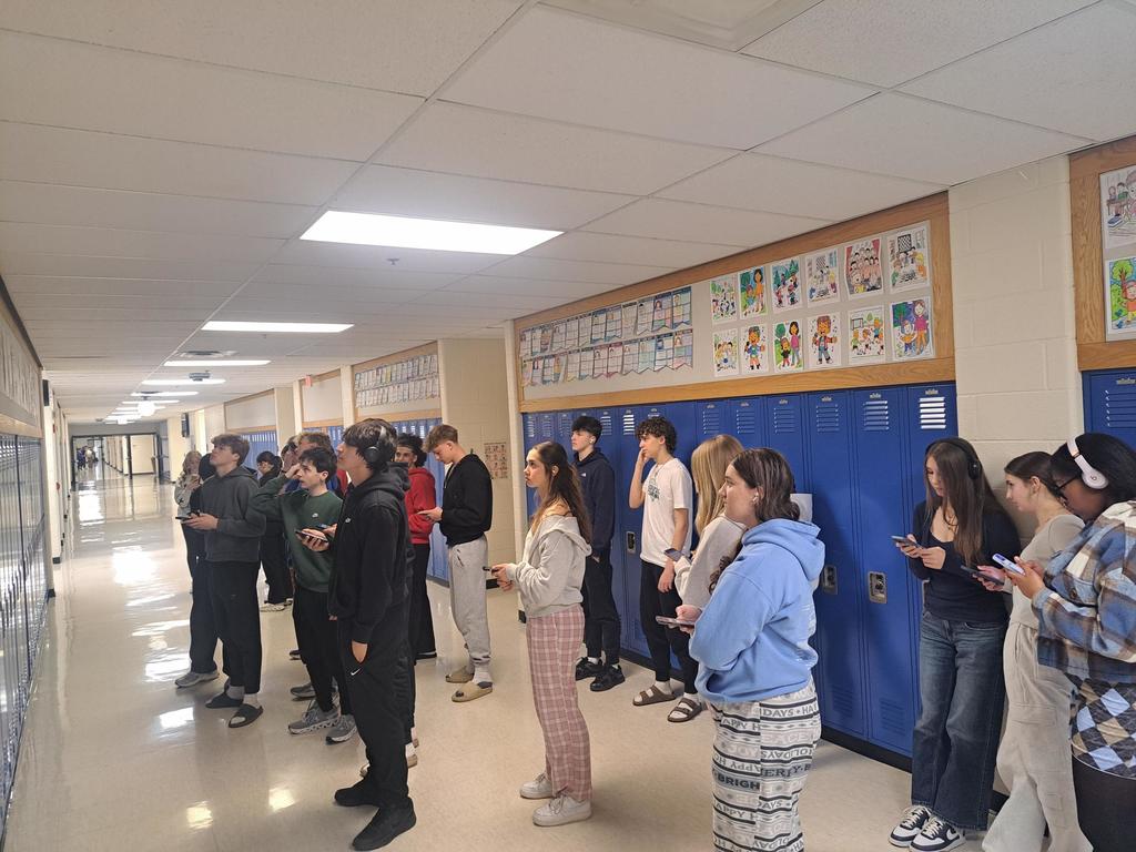People stand in a hallway, facing a blue wall with posters. Some wear hoodies, sneakers, and headphones.