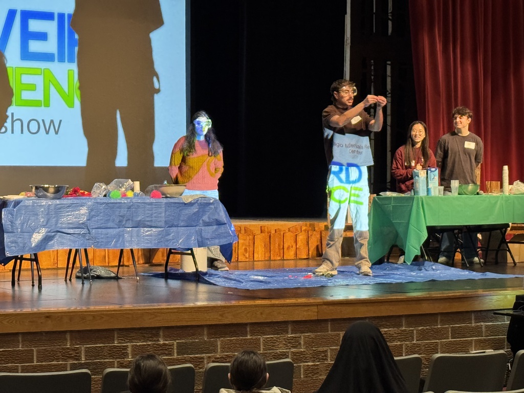 Four people stand on a stage with tables in front of an audience. One person wears a blue face paint.