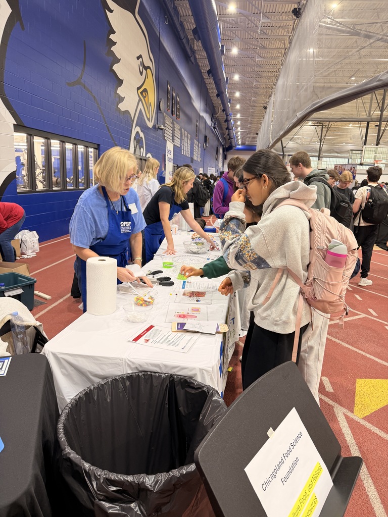 People at tables inside an indoor sports facility. Tables have papers, pens, and trash bins.