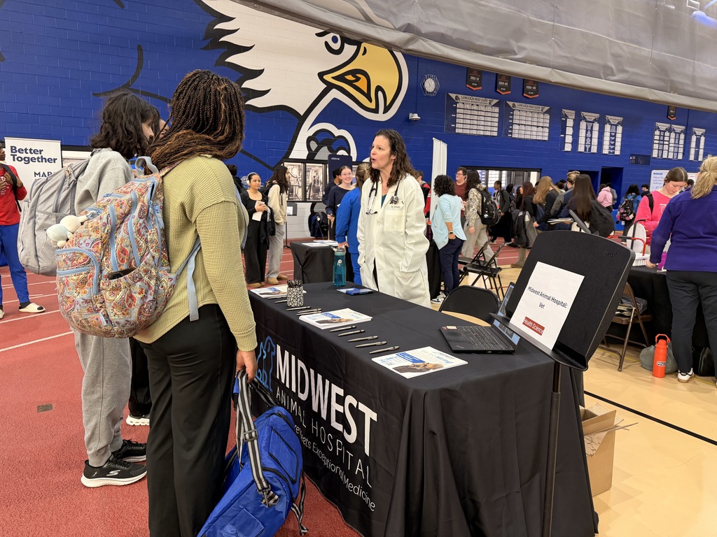 A gym with a table has "Midwest" and "Hospital" on the tablecloth. Several people stand and talk.