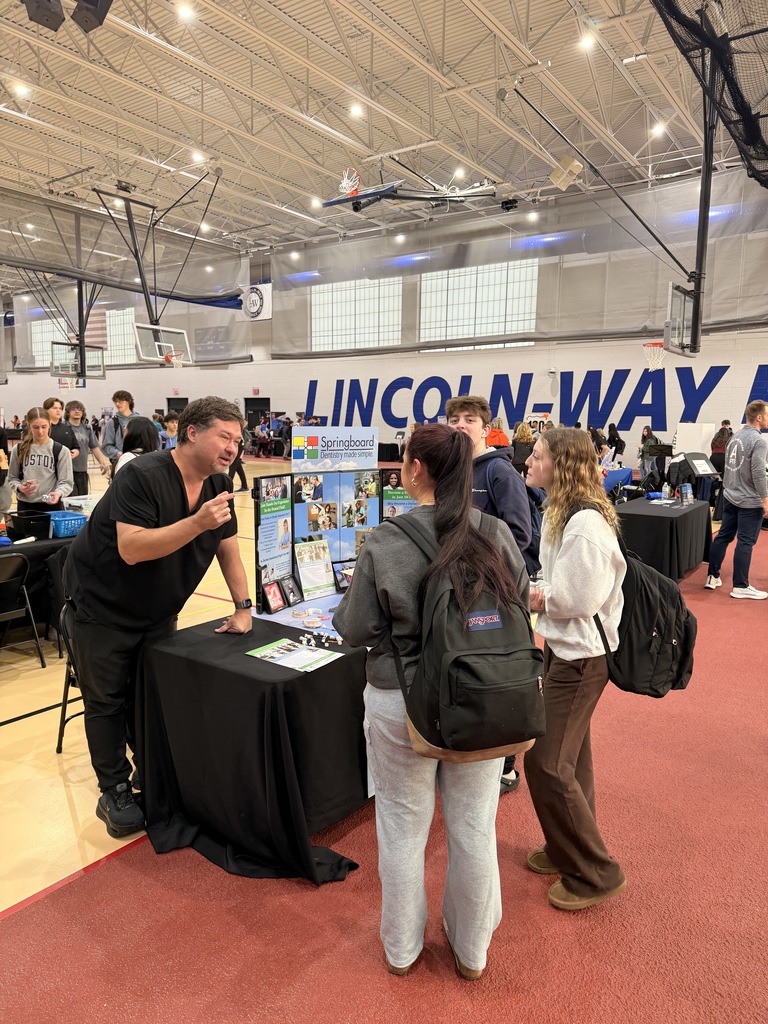 People in a gym, one man speaks at a table with a black cloth, three women stand nearby.