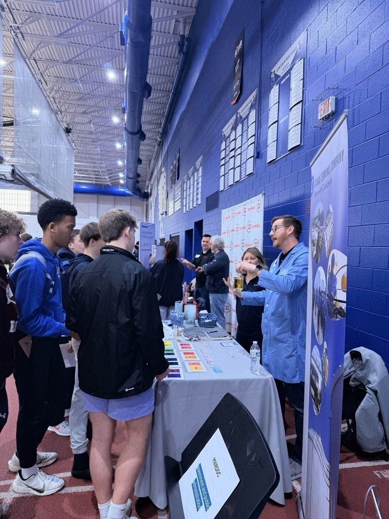 Indoor area with blue walls. People around a table with items. One person in a lab coat explains something.