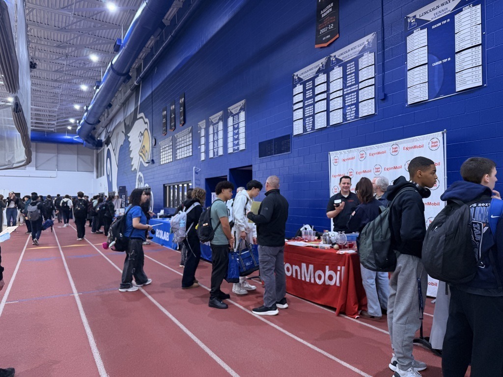 People line up in a gym, with blue walls and red track, to speak with people at a table.