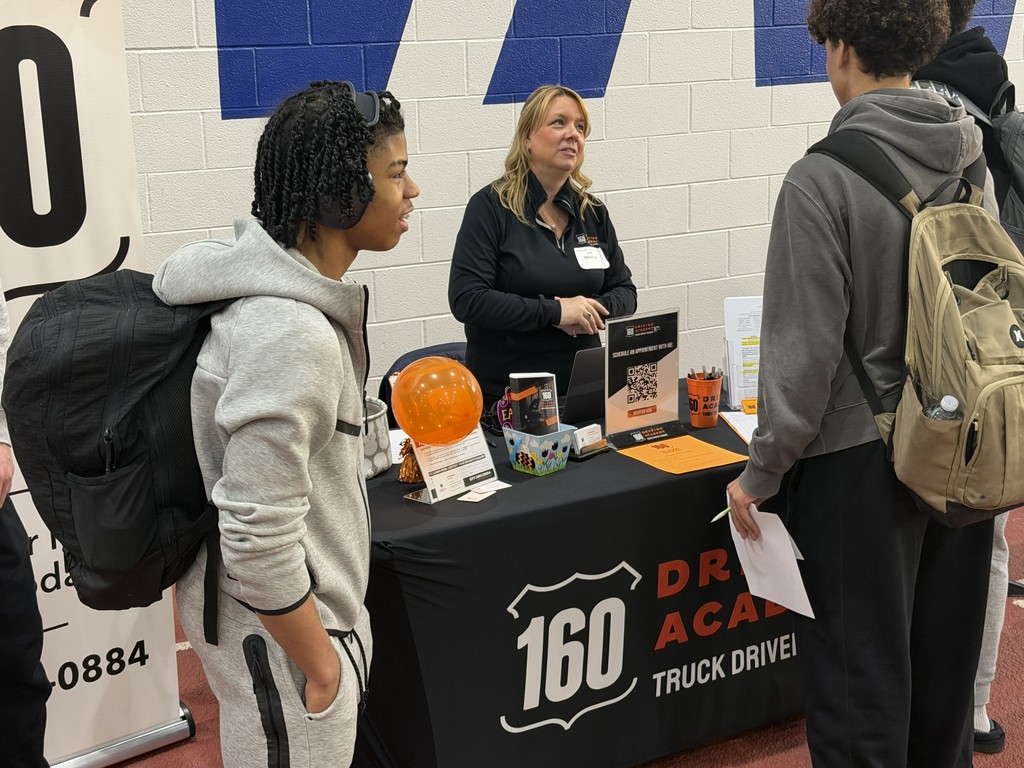 People stand at a table with a black tablecloth and sign reading "DR 60 ACA Truck Drive!" Two wear backpacks.