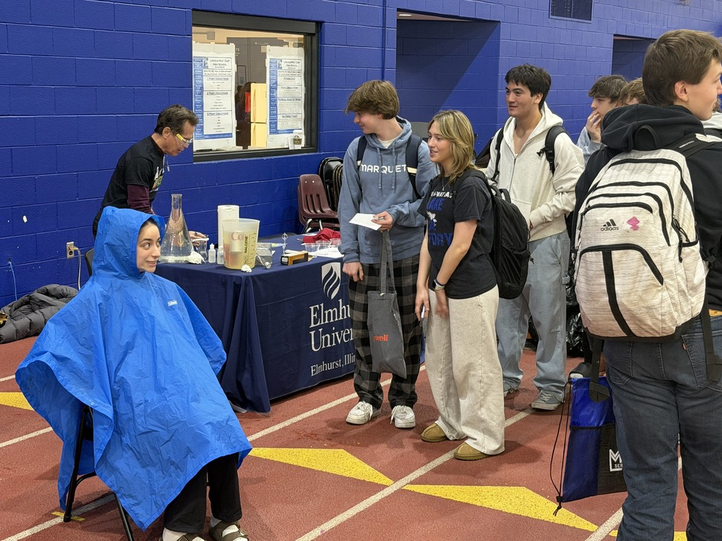 People stand near a table and track with a blue backdrop. One person sits in a chair wearing a blue poncho.