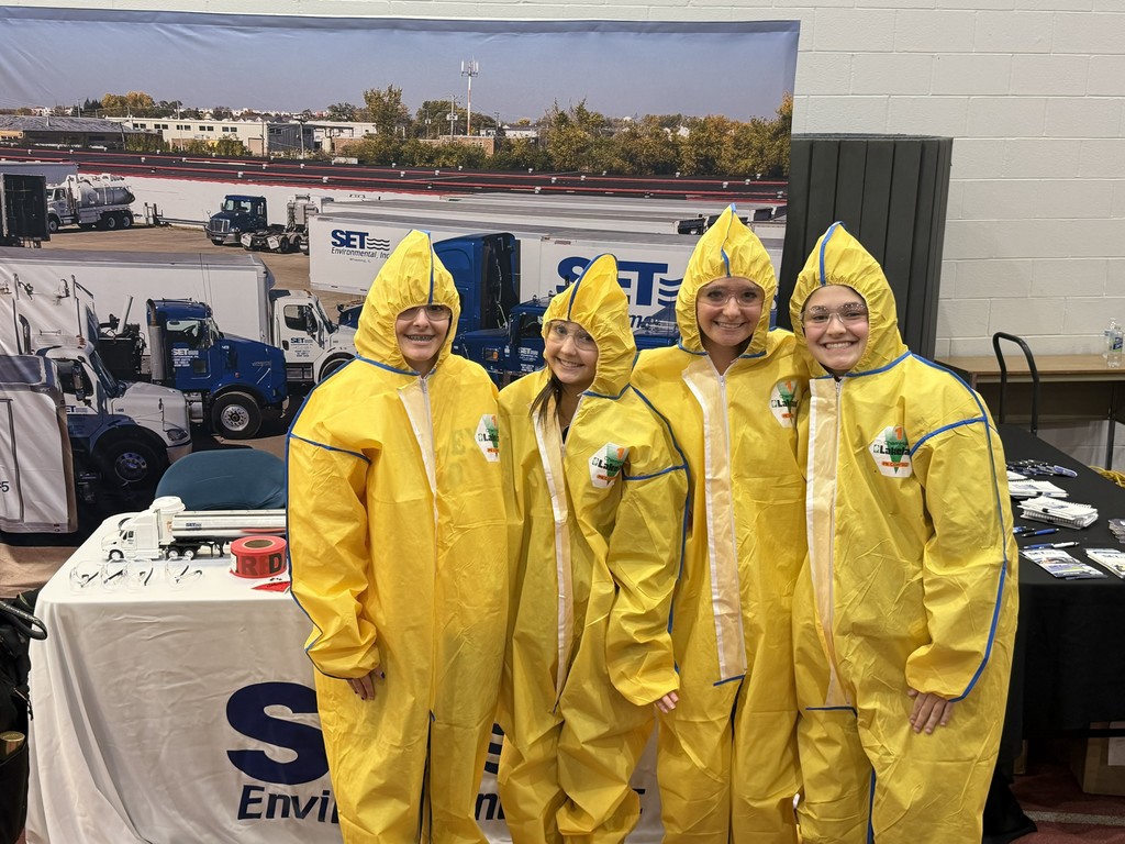 Four people in yellow protective suits stand together in front of a table. A white banner reads "S Env"