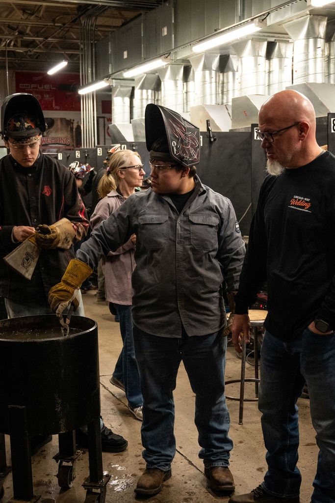 Three men in a workshop. One holds a metal container, another wears a welding helmet, and the third wears glasses.