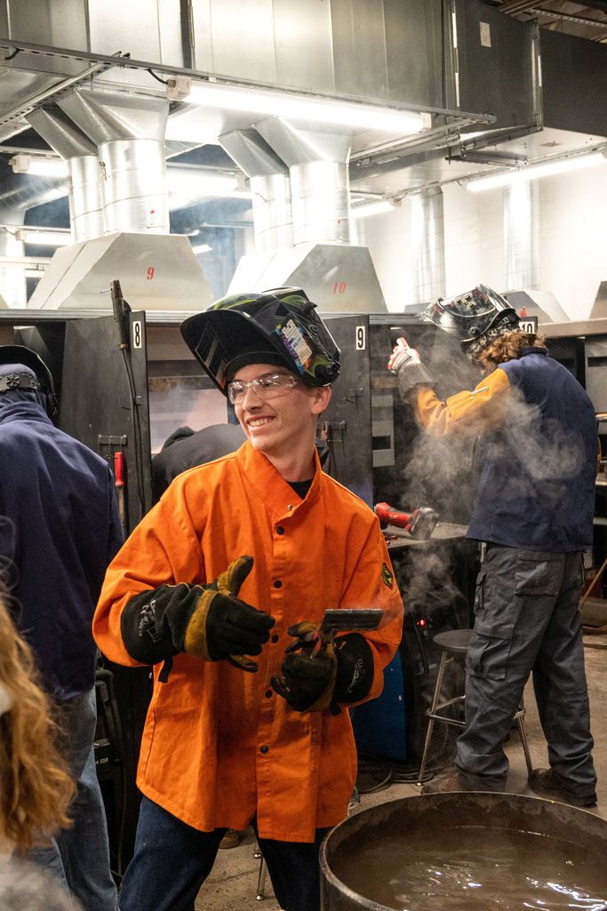 Individuals in orange coats and helmets stand in a workshop. One holds a tool, another works at a machine.
