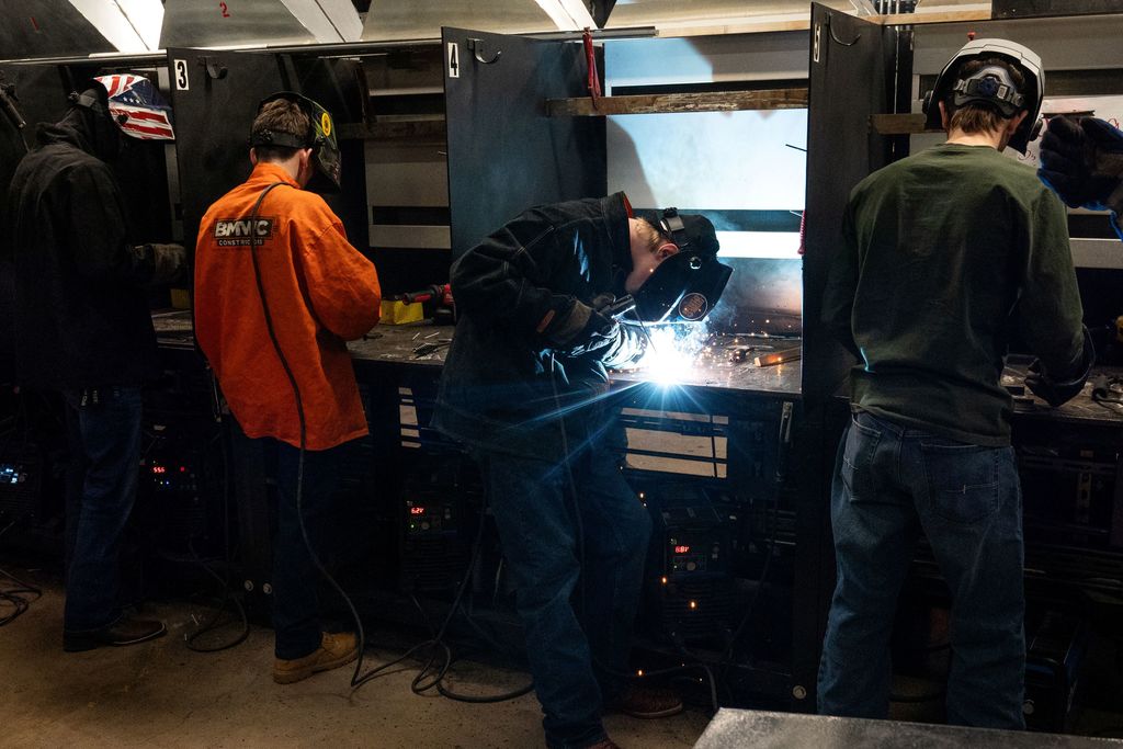 A room with a workshop setup. Several men wearing safety gear work on metal projects. One man is welding.