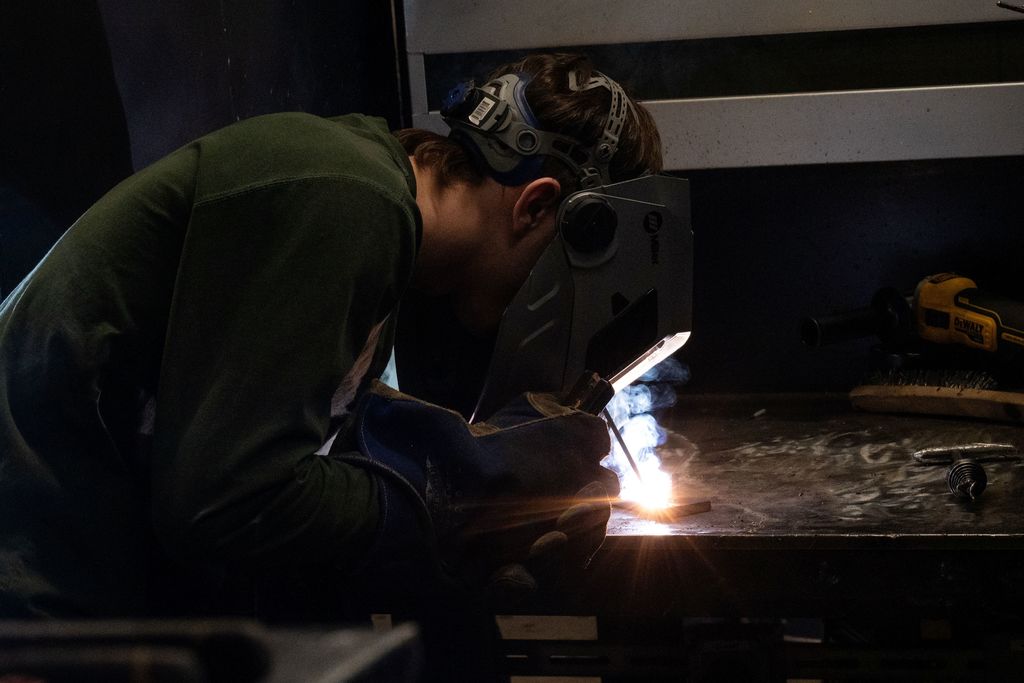 A person in a green shirt and welding mask works on a metal table with a welding torch.