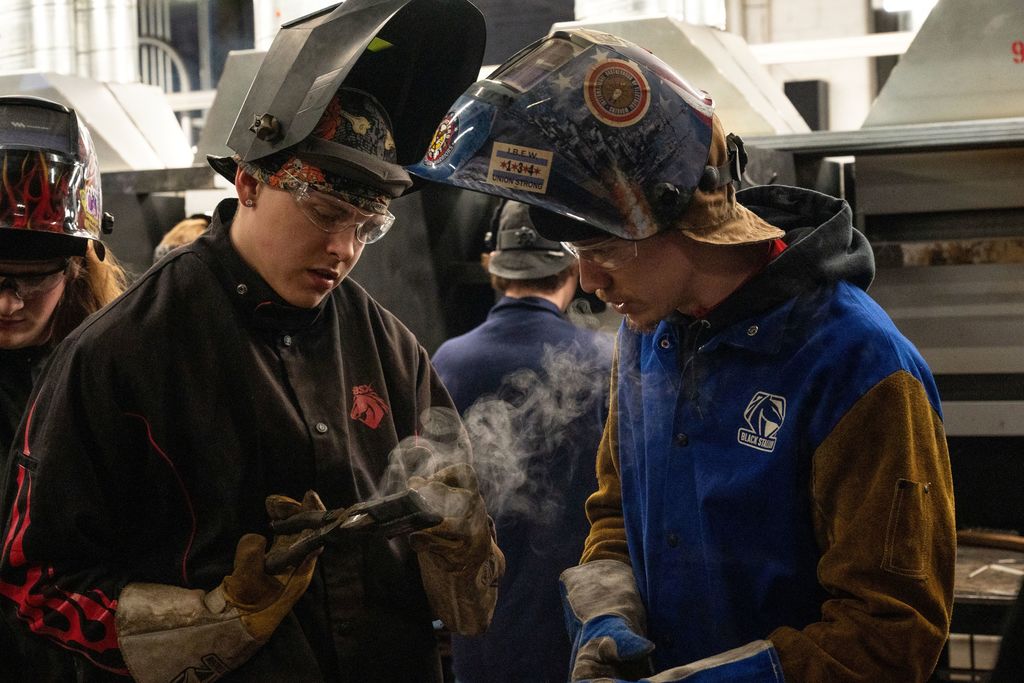 Two people wearing safety helmets and gloves work together in a workshop, one holding a welding mask.