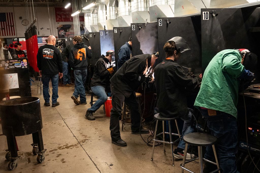 People wearing protective gear in a workshop; some use welding torches and others stand in line.