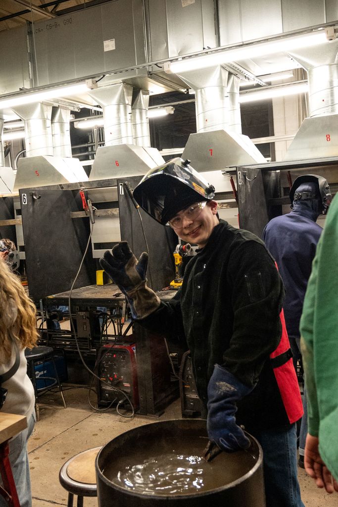 A person stands in a workshop with a protective helmet, smiling and holding a metal bowl.
