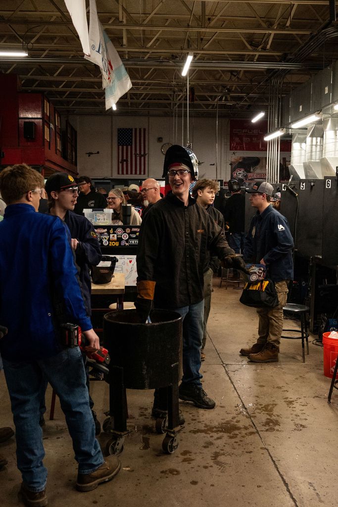 A workshop with metalworking equipment. People gather around a man who stands before a bucket.