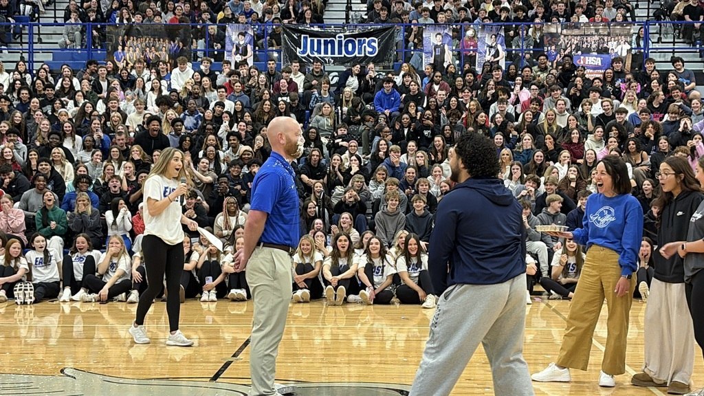 People gathered on a basketball court, some holding a bat. One person stands at a microphone.