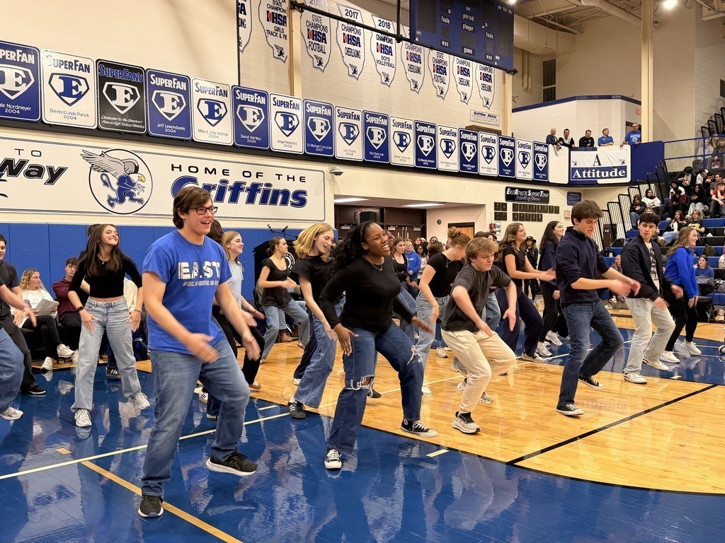 A group of people dance in a gym. They wear various colored shirts and sneakers. The gym has a wooden floor and banners.