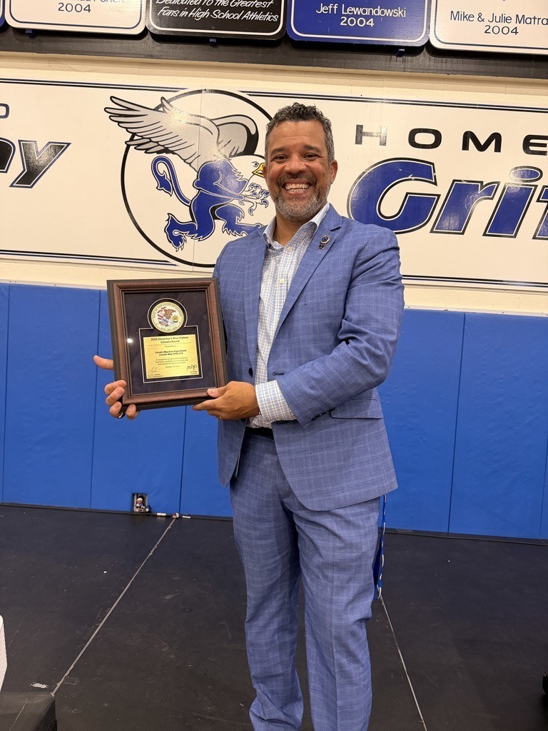 Mr. Griggs  in blue suit holding a framed award in a gym with a blue and white wall.