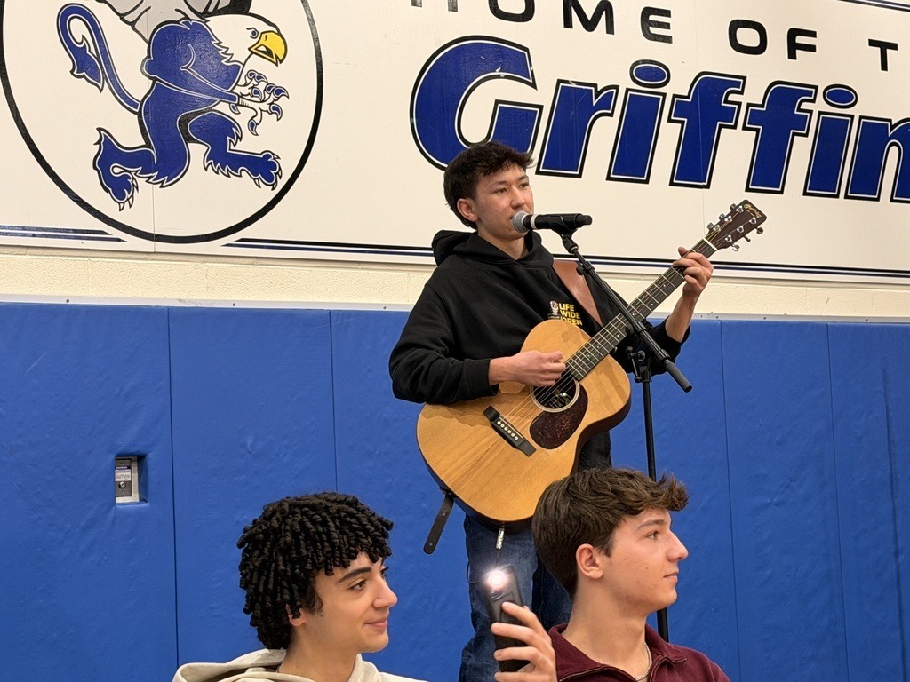 A student plays guitar and sings into a microphone. Two young people look on from below. Behind him is a banner reading "Home of the Griffins."