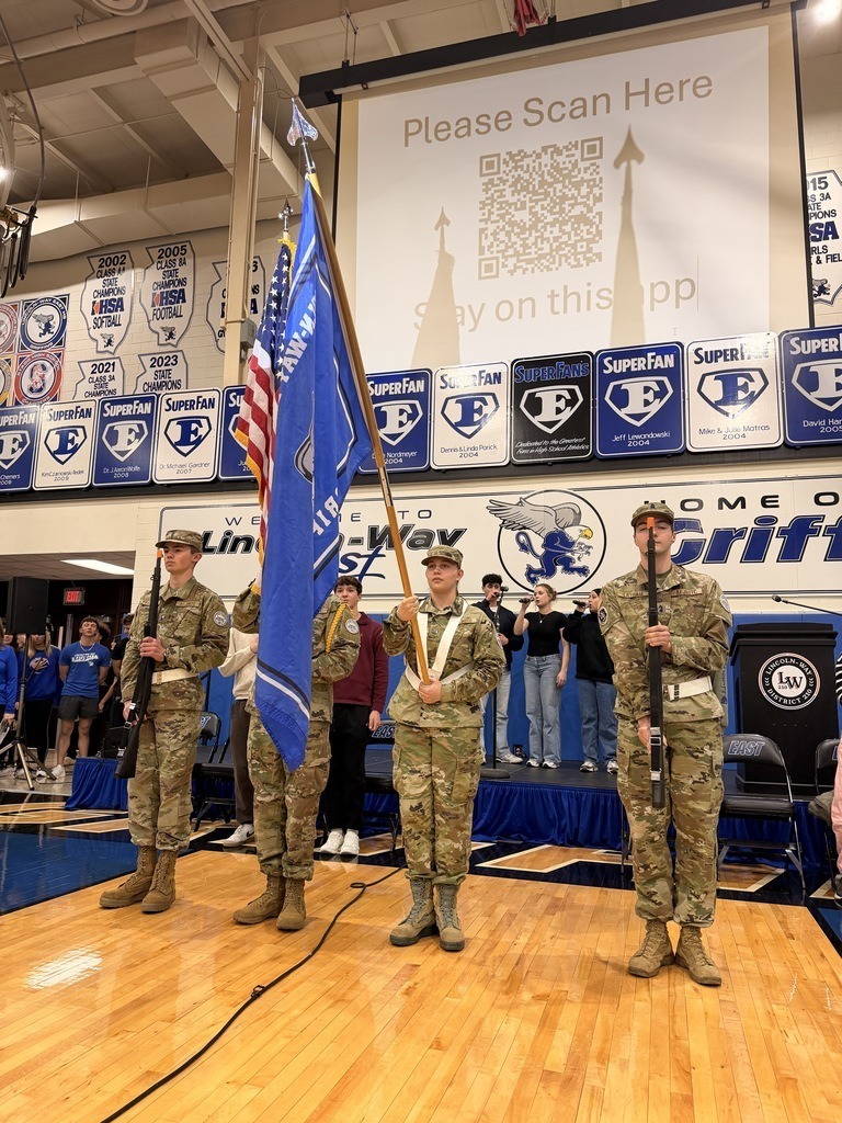 Four military personnel in uniform stand on a stage with a flag. A large screen in the background displays a QR code.