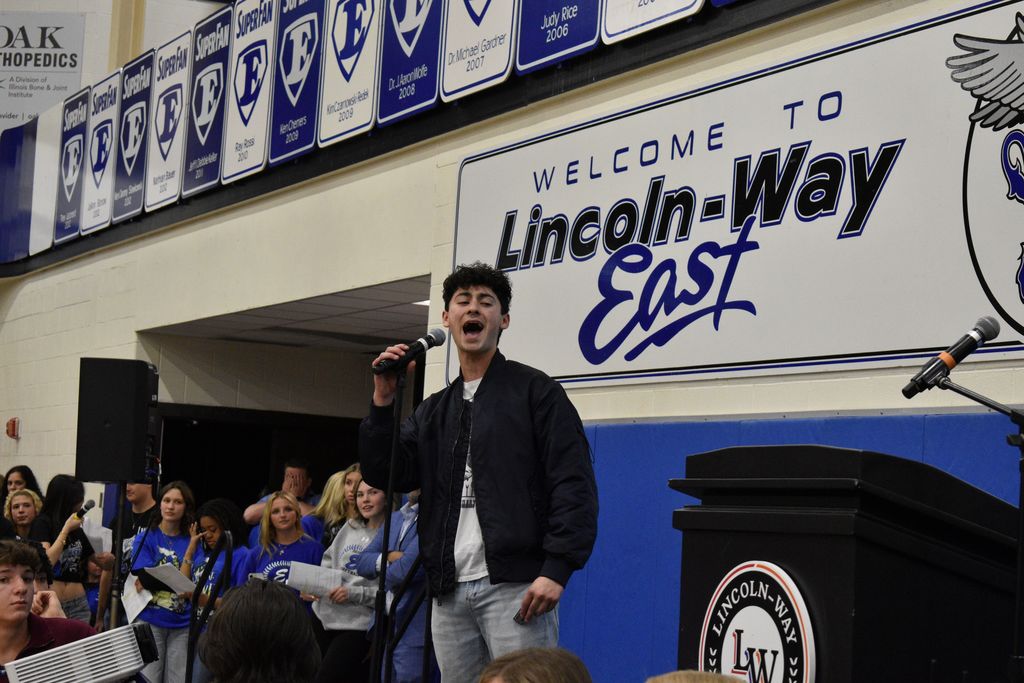 Person singing into a microphone in a room with a blue wall. A sign reads "Welcome to Lincoln-Way East."