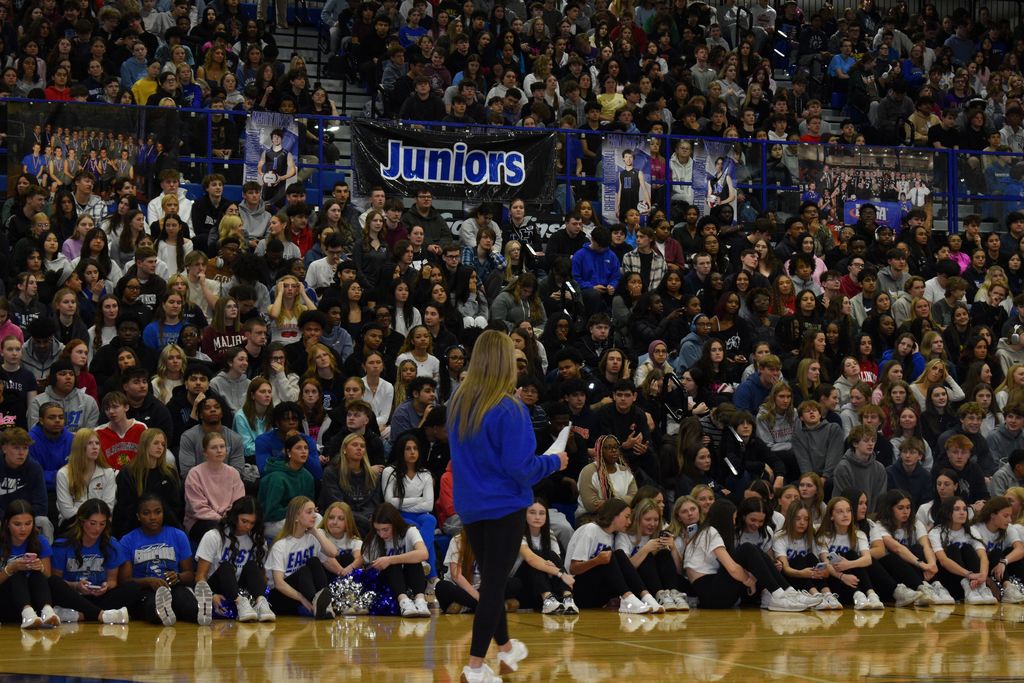 A large audience of people, mostly students, fills the bleachers of a basketball court. A student stands in front, holding a paper.