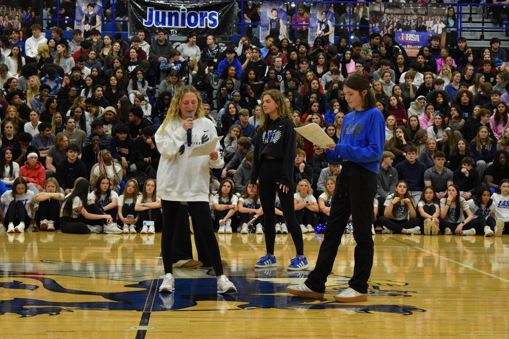 Three students on a basketball court with a crowd. One holds a microphone, one reads a paper, and one watches.