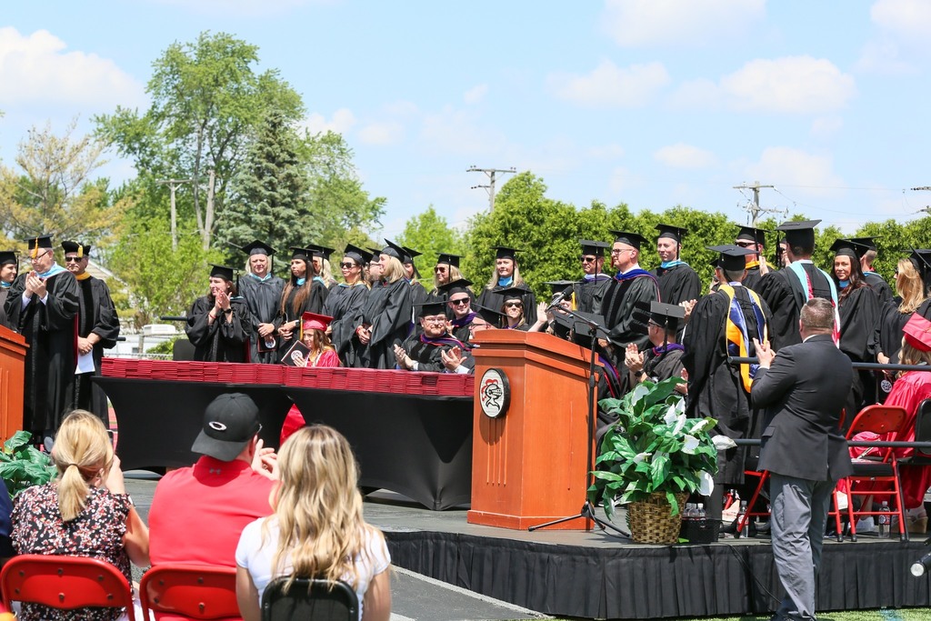 Graduates in gowns and caps stand in line on stage, with a podium and green plant, while spectators sit in chairs.