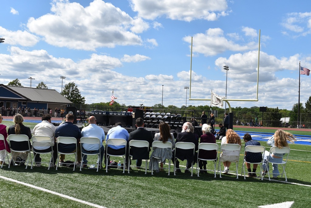 A group of people sit in chairs on a football field, with a stage and flags in the background.