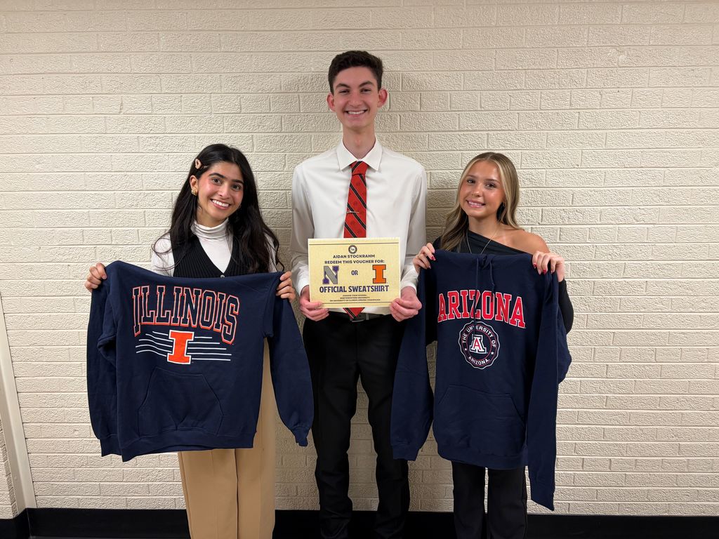Three people pose for a photo; two hold blue sweatshirts, one holds a certificate. White brick wall in the background.
