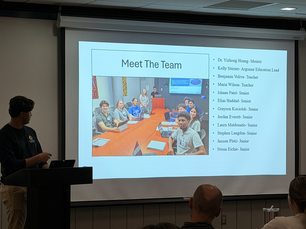 A person stands at a podium in front of a projector screen with a slide titled "Meet The Team".