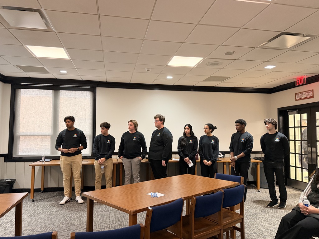 A group of individuals in black shirts and pants stand in a room with tables and chairs.