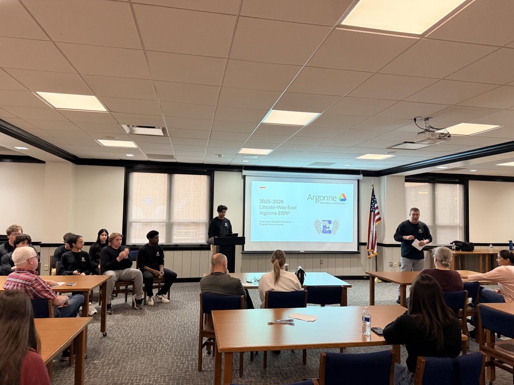 Classroom with tables and chairs, people sitting, a man stands at a podium in front of a projector screen.