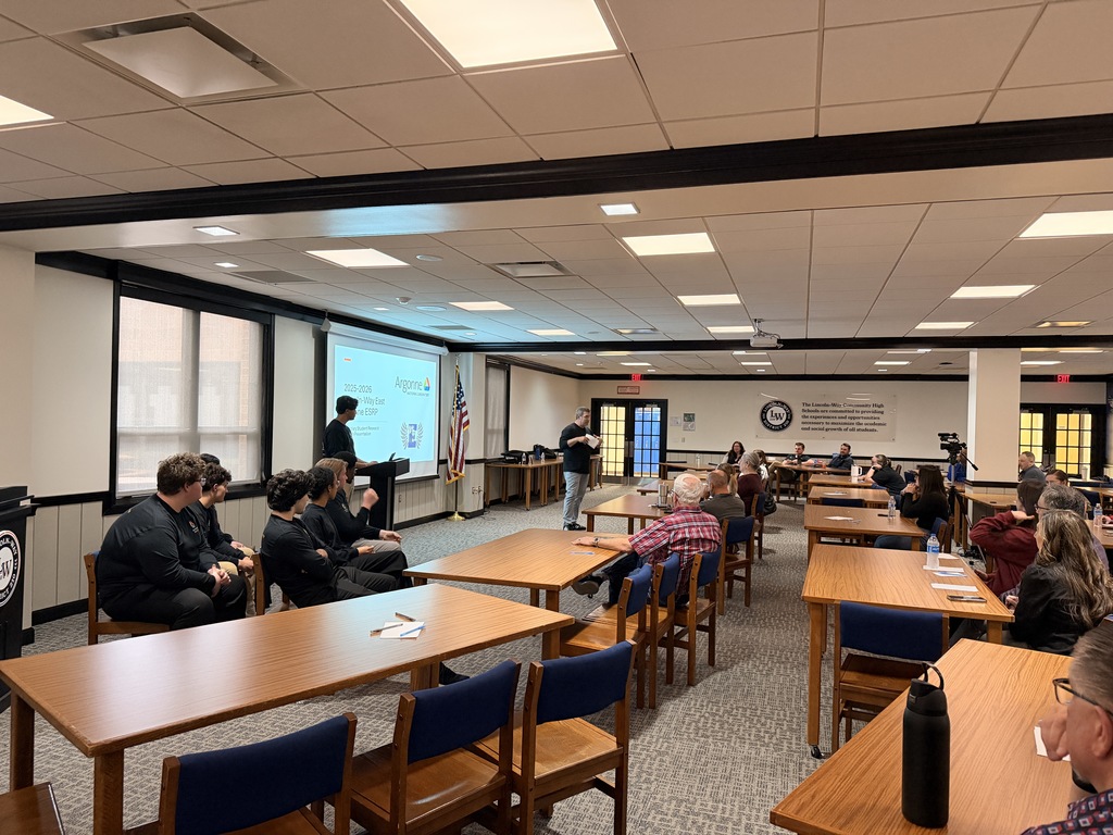 A room with tables and chairs arranged in rows, with a person speaking in front of a projector screen.
