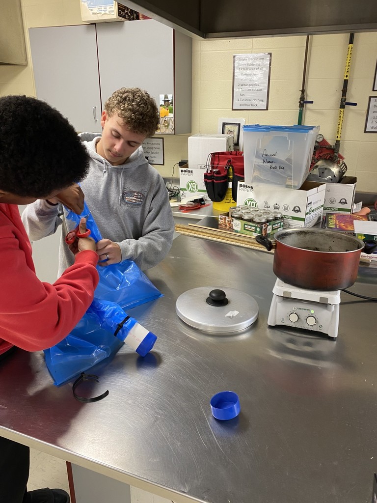 Two students work in a kitchen, one handling a plastic bag and the other preparing an item on a hot plate.