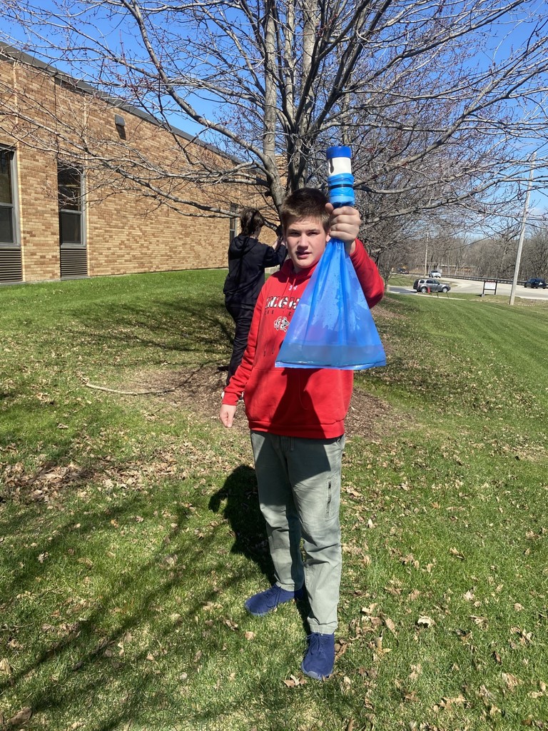 A person stands in grass holding a blue bag and a blue bottle. Behind them, a tree with bare branches.