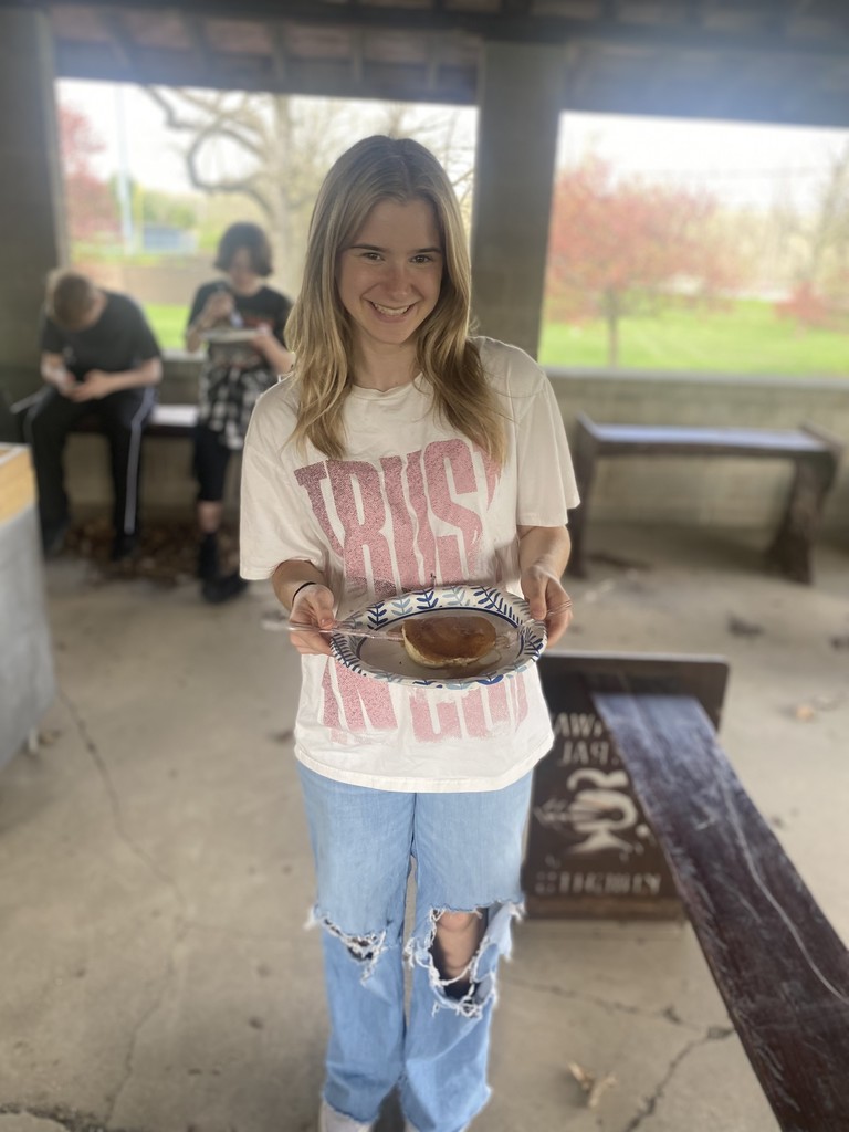 A student smiles while holding a plate with food, wearing a T-shirt and ripped jeans. Two people sit behind her.