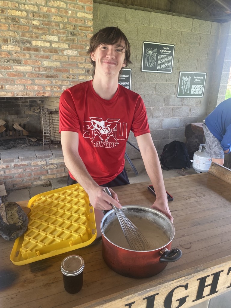 A student in a red shirt stirs batter in a pot using a whisk. Behind him, a fireplace and two yellow trays are visible.