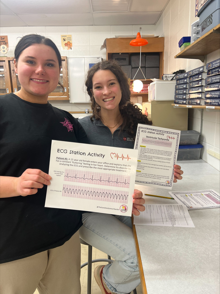 Two women sit together, one holds a paper with a diagram of an EKG. They both smile at the camera.