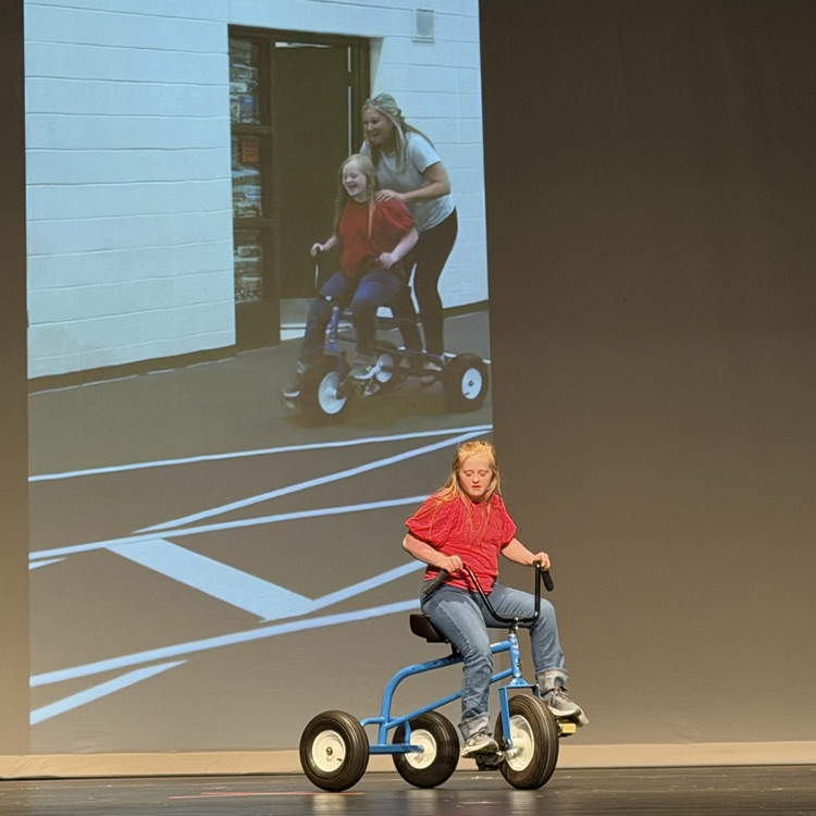student riding a tricycle on stage with a photo of them as the background