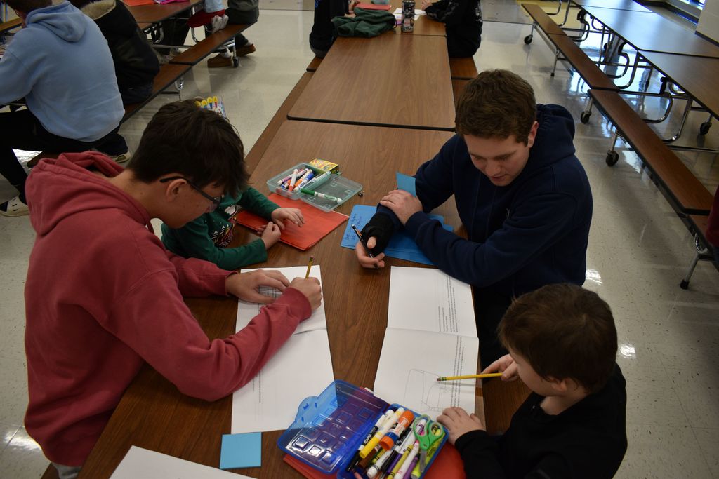 Three boys in hoodies work at a table, with papers and coloring tools. Behind them, students sit at tables.