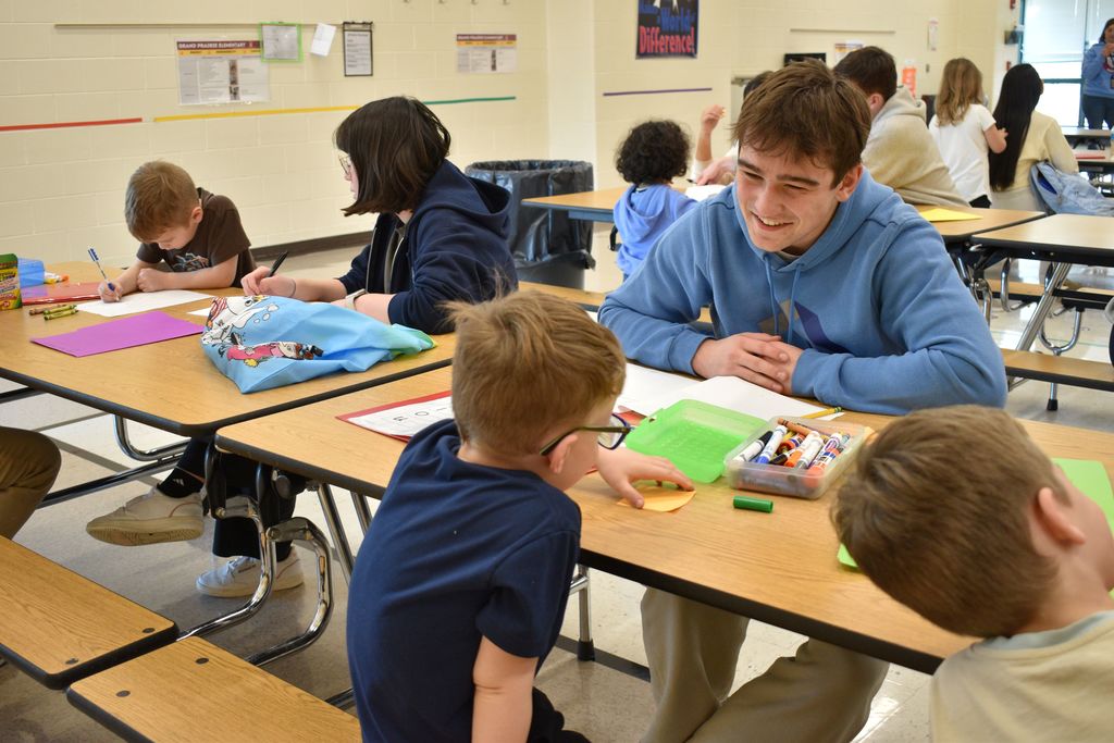 Several children sit at tables, engaging in coloring activities. A high school student in a blue hoodie stands nearby, assisting a child.