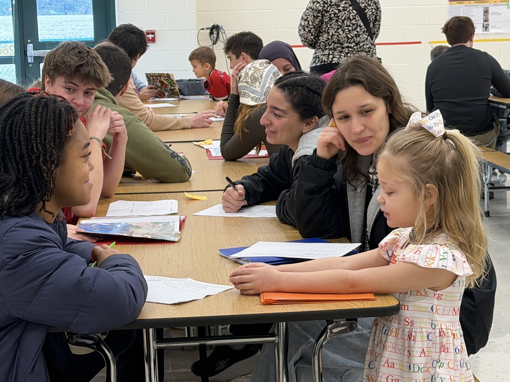 Several students sit at desks, with a high school student and girl in front, working on papers and books.