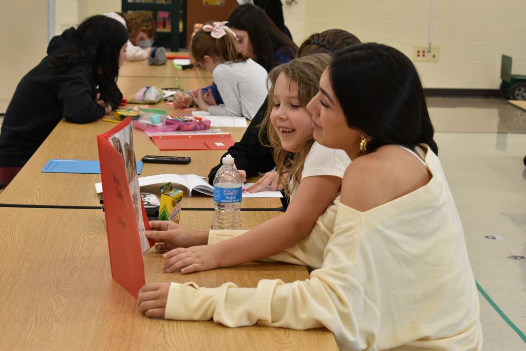 Group of students in a classroom. One student is smiling, another holds a red folder. Books, pens, and a water bottle on the table.