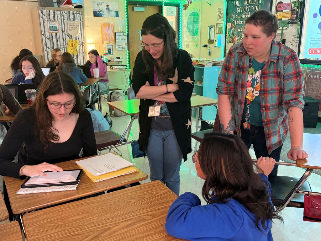 Four individuals in a classroom with a teacher at a desk. Two students sit at desks, one using a tablet.