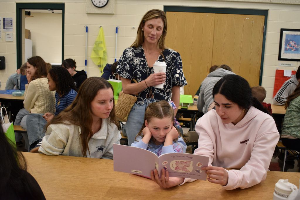 A woman with a cup stands behind three girls reading a book at a table. Other students sit at tables in the background.