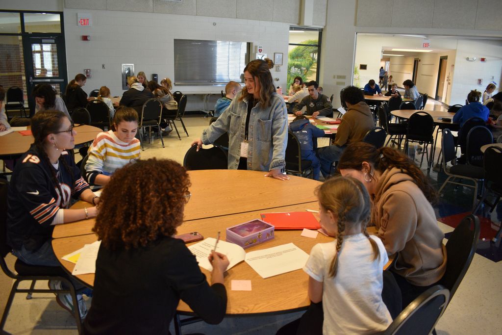 Group of adults and children sitting around a table in a classroom. Papers and writing utensils on table.