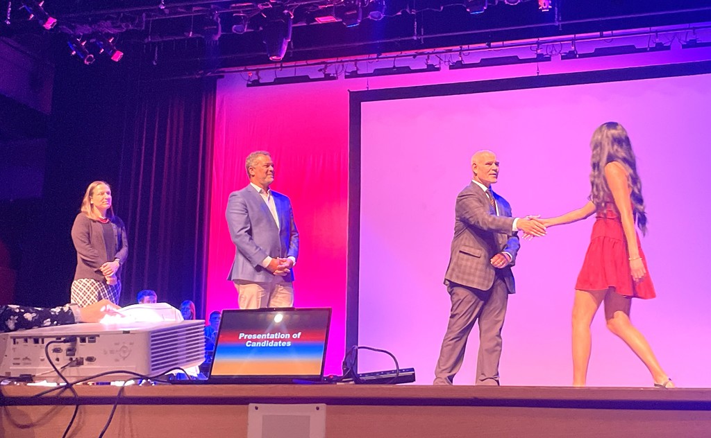 Three people on a stage, one in a red dress shaking hands with a man in a suit.